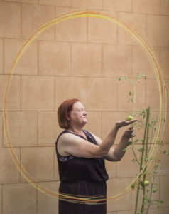 A photo of a woman behind a window. She is holding a tomato growing on a plant.