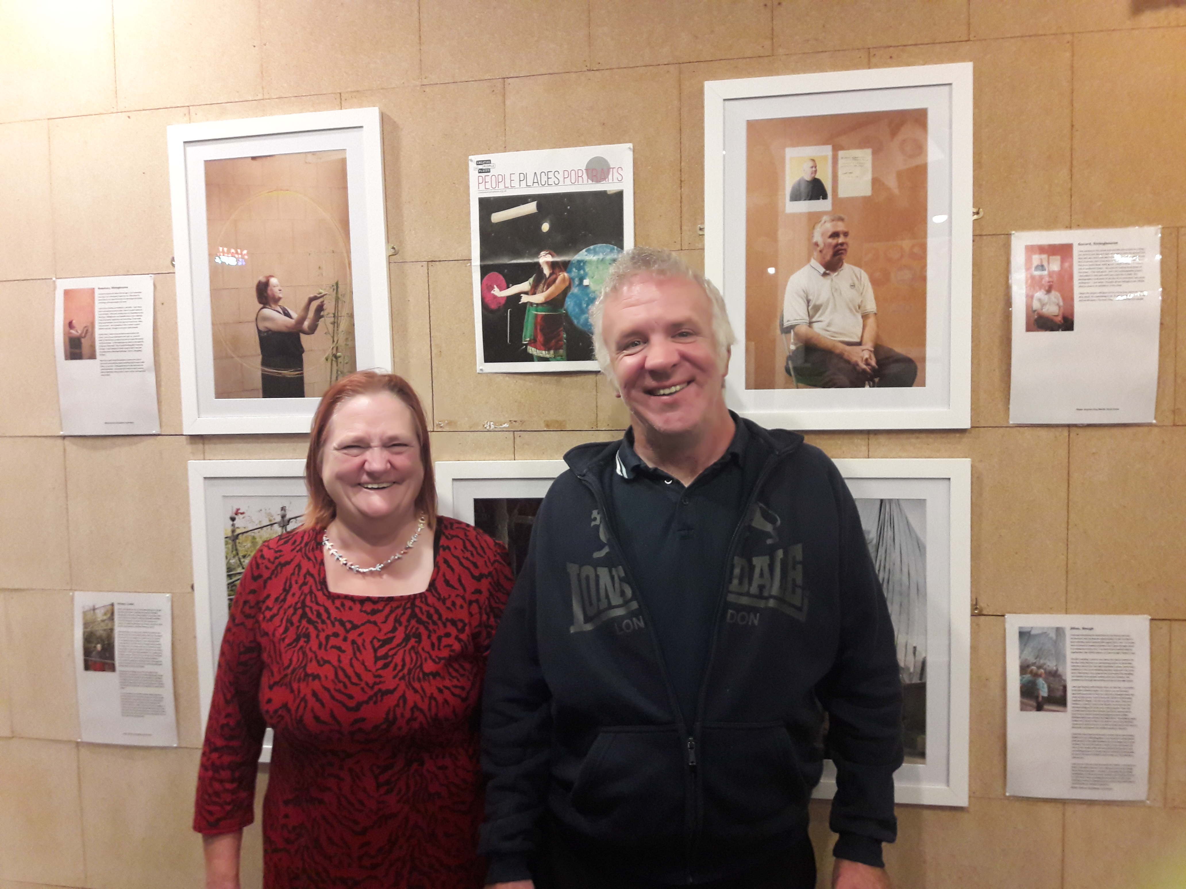 Two people stand in front of an exhibition which contains portraits of them.