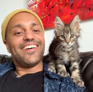 Anil is a smiling person who is wearing a yellow beanie hat and a blue shirt with a black top underneath. They are sitting on a black sofa and have a red artwork behind them. On Anil's shoulder is a very fluffy grey tabby cat who is looking at the camera.