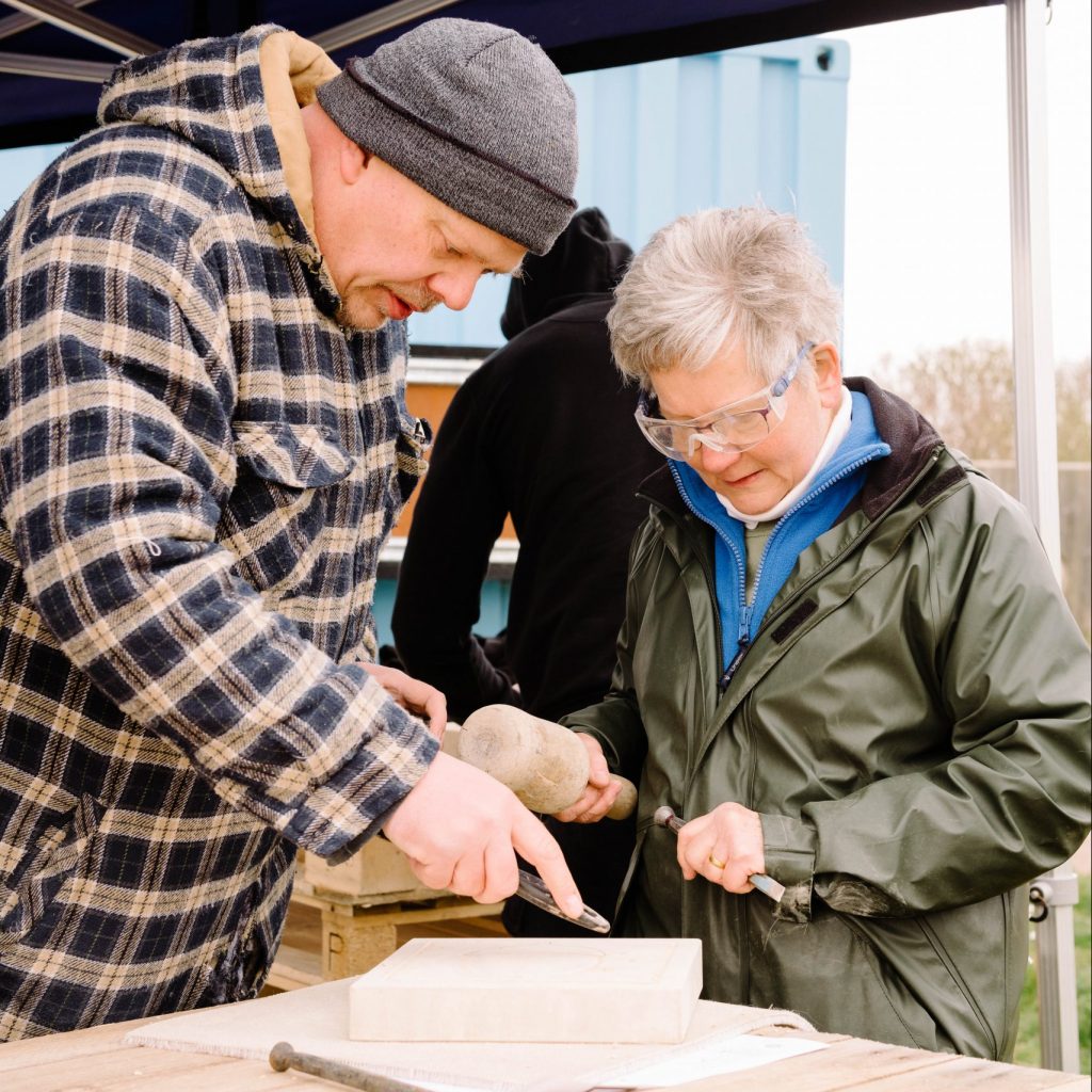 Patrick, a tall white man, is giving stone carving advice to Lynda from The Friends Of Milton Creek Country Park. Lynda is a white woman with grey hair and is wearing safety glasses