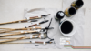 A range of paintbrushes, taped together in pairs, are laying down facing right, dipped in ink. Next to them are 3 pots of dark liquid.