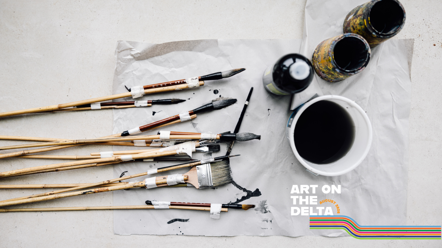 A range of paintbrushes, taped together in pairs, are laying down facing right, dipped in ink. Next to them are 3 pots of dark liquid.