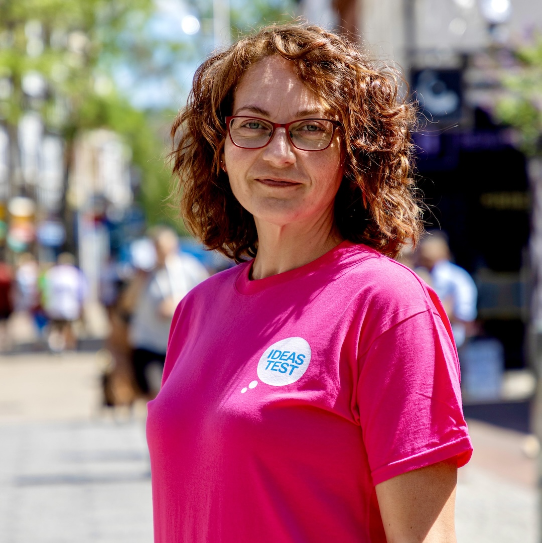 A woman with auburn curly hair, wearing glasses and a bright pink t-shirts, stands outside and smiles at the camera
