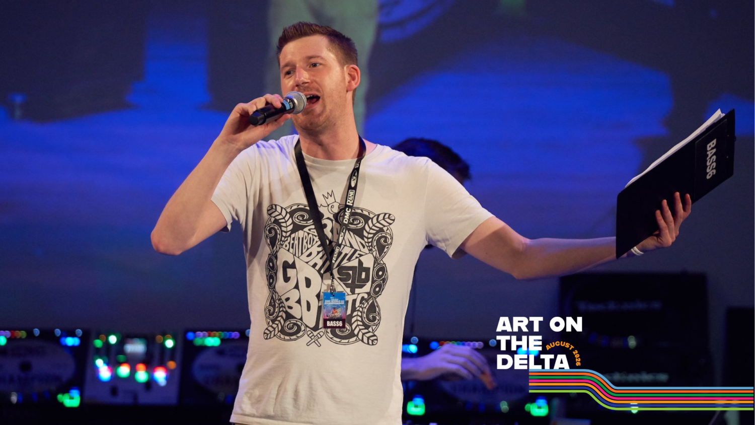 A white man in his mid-30s stands in front of a blue screen with a mic, and holding a clipboard in his other hand