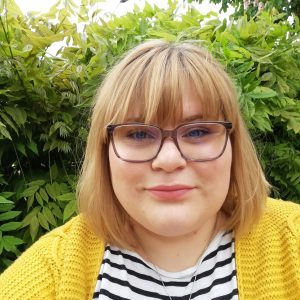 Bryony is a white woman with a light brown bob haircut with blonde highlights. She is wearing glasses, a yellow cardigan and a top with black and white horizontal stripes. She has a smile on her face and is standing outdoors in front of a green bush.